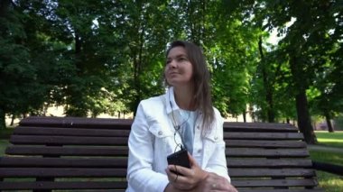 Beyaz kadın telefonda kulaklıkla şarkı söylüyor. Parktaki bankta oturuyor.