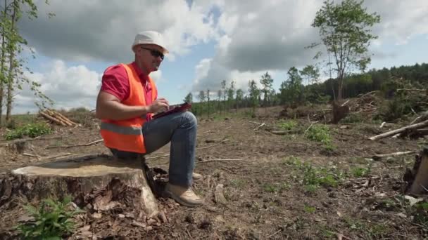Мужчина в шлеме сидит на пне лесозаготовительной территории — стоковое видео