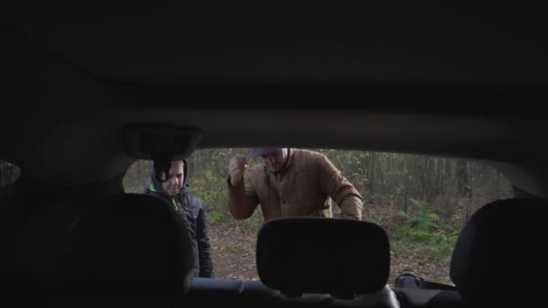 Отец и сын, открывая багажник машины, берут рюкзак и идут в лес. — стоковое видео