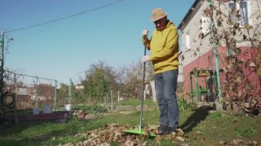 Şapka ve eldiven giyen kıdemli çiftçi kurumuş yaprakları süpürüyor.