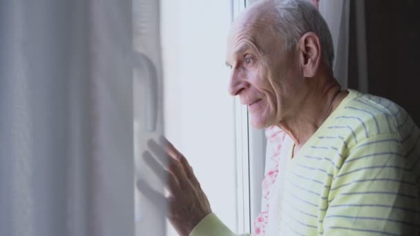 Glimlachende oudere man kijkend door vensterglas en zwaaiend groet gebaar. — Stockvideo