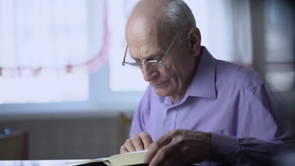 Idős, ősz hajú, szemüveges férfi felolvassa a hangos könyvet, miközben az asztalnál ül. — Stock videók
