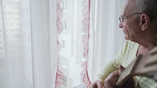 Alter Mann mit Brille spielt Akustikgitarre und blickt durch Fenster. — Stockvideo