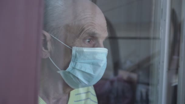 Vista lateral cerrada del anciano en la máscara de cara mirando detrás del vidrio de la ventana. — Vídeos de Stock