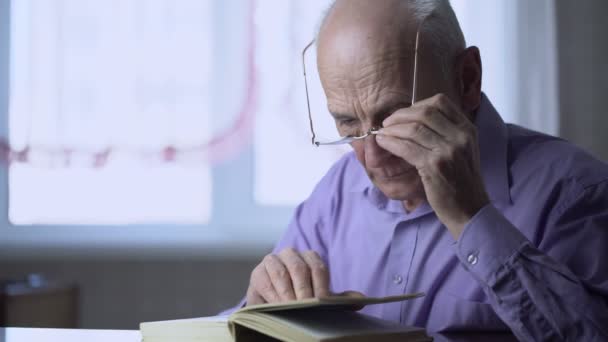 Homem de cabelos grisalhos solitários com óculos lendo livro sentado à mesa — Vídeo de Stock