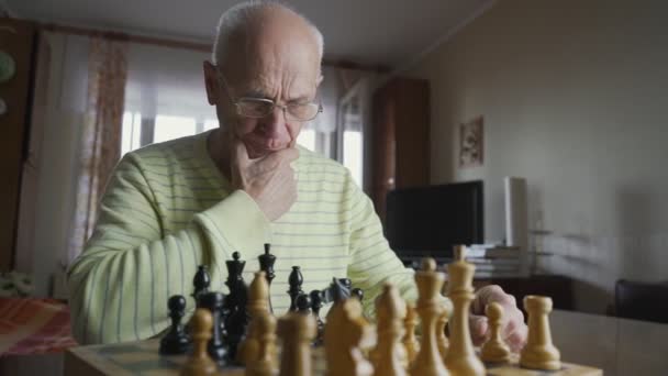 Äldre man med glasögon spelar schack med sig själv när han sitter vid bordet — Stockvideo