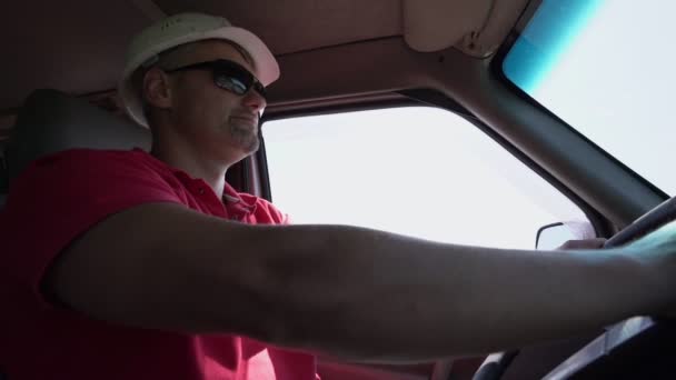Человек с бородой в строительном шлеме водит машину — стоковое видео