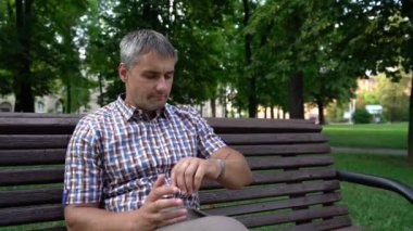 Adam etrafına bakıyor ve zamanı kontrol ediyor. Bekliyor ve parkta oturuyor.