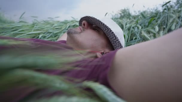 Mannelijke boer in hoed liggend op tarweveld luisteren naar muziek in hoofdtelefoon — Stockvideo