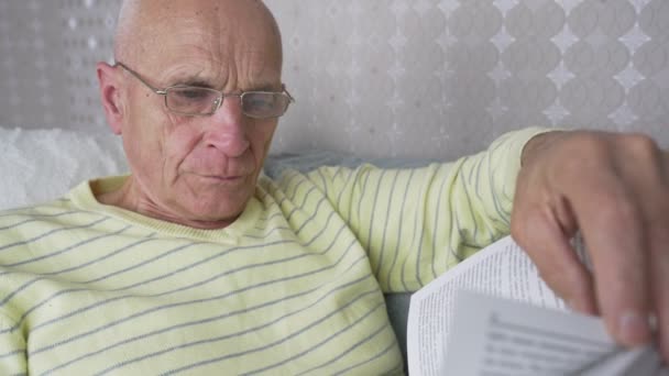 Зрелый мужчина в очках читает книгу, лежащую на диване — стоковое видео