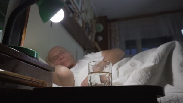 Älterer Mann liegt im Bett unter weißer Decke und Glas Wasser auf Nachttisch. — Stockvideo