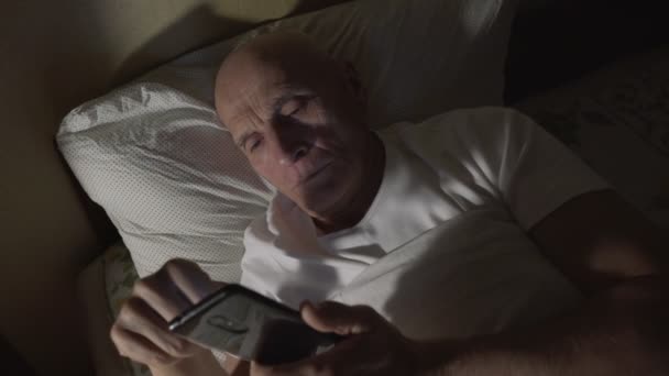 Rentner mit faltigem Gesicht liegt nachts mit Smartphone im Bett — Stockvideo