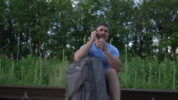 Homem com mala sentado em trilhos na estrada de ferro e falando ao telefone — Vídeo de Stock