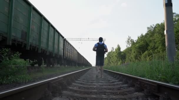 Чоловік мандрівник з рюкзаком прогулянки далеко вздовж рейок залізниці біля прохідного поїзда — стокове відео
