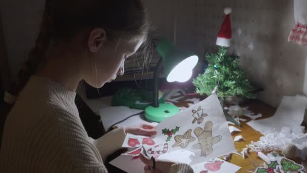 Genç kız masada oturup Noel Baba 'nın şapkasını kağıt çarşaftan kesiyor. — Stok video