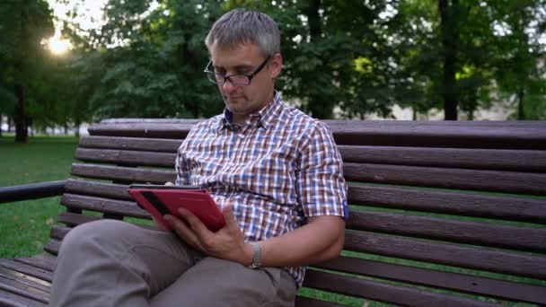 Hombre con anteojos trabaja usando tableta sentada en el parque de la ciudad — Vídeos de Stock