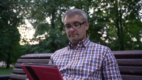 El hombre trabaja en la tableta bebiendo café de la taza de papel en el parque — Vídeos de Stock