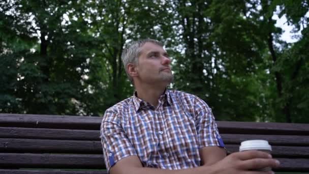 男人坐在公园的长椅上，一边喝着纸杯里的咖啡 — 图库视频影像