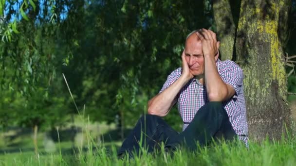 Плачущий взрослый мужчина в рубашке держит голову, опираясь на дерево — стоковое видео