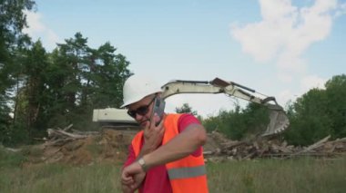 Foreman kask ve yelek giyip telefonla konuşuyor ve saati kontrol ediyor.