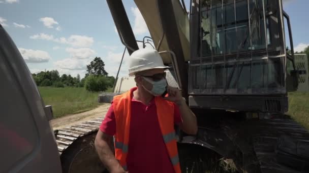 Foreman in medical mask stands near excavator gives instructions by phone — Video Stock