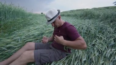Şapkalı adam buğday tarlasında oturmuş buğdayın üstüne kulaklık takıyor.