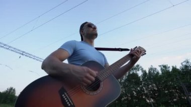 Açık havada durup gitar çalan güneş gözlüğü takan bir müzisyen.