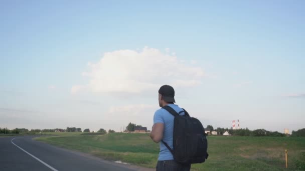 Hombre con gorra y mochila camina a lo largo del centro de la carretera cerca del pueblo — Vídeos de Stock