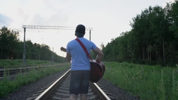 Homem caminha ao longo de trilhos de trem e toca guitarra no dia de verão visão traseira — Vídeo de Stock