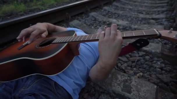 Людина в сонцезахисних окулярах лежить на шпалах залізниці грає на гітарі — стокове відео