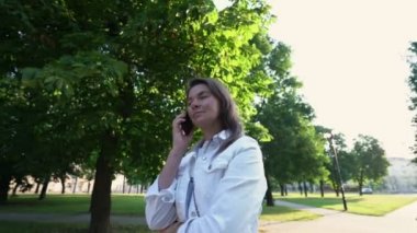 Beyaz ceketli kadın güneşli parkta telefonla konuşuyor.