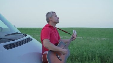 Adam akustik gitar çalıyor ve minübüs kaputunun önünde şarkı söylüyor.