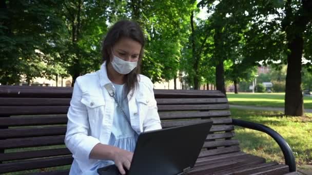 Mulher em tipos de máscara no laptop sentado no banco do parque — Vídeo de Stock