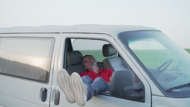 El hombre duerme en el asiento delantero de la minivan inclinando sus piernas por la ventana — Vídeo de stock