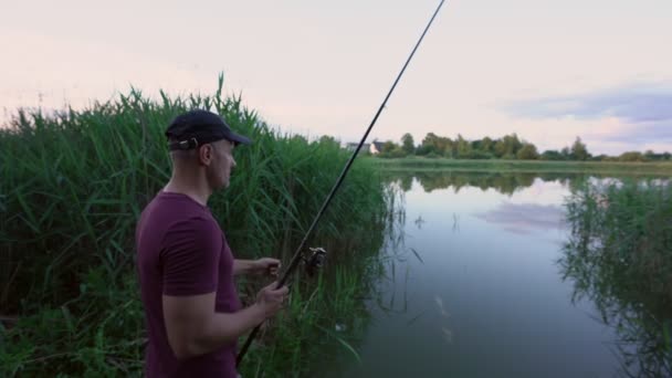 Άνδρας ψαράς με καπέλο ρίχνει καλάμι ψαρέματος από την ακτή σε μικρή λίμνη — Αρχείο Βίντεο