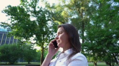 Beyaz elbiseli esmer kadın parkta ayakta duruyor ve telefonla konuşuyor.