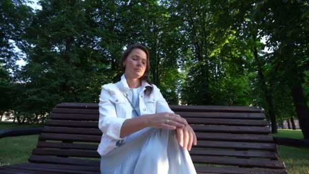 Middle aged woman in white sits waiting on shadow bench in park — Stockvideo