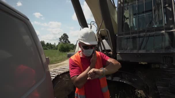 Bauarbeiter mit Mundschutz, orangefarbener Weste und weißem Helm telefoniert — Stockvideo