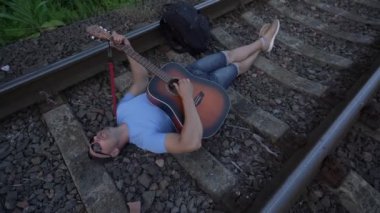 Güneş gözlüklü ve şortlu bir adam demiryolu yatağında yatıyor ve gitar çalıyor.
