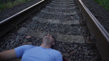 Güneş gözlüklü bir adam demiryolunda yatarken yukarı bakar ve gitar çalar.