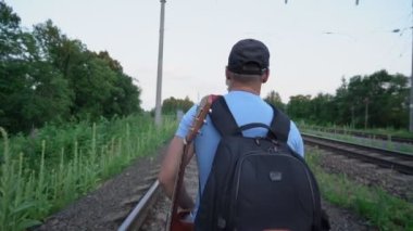 Adam demiryolunda yürüyor, sırt çantası ve gitar taşıyor.