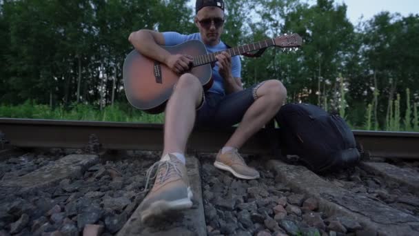 Demiryolu raylarında oturan adam gitar çalıyor, parmaklarını tellerin üzerinden geçiriyor. — Stok video