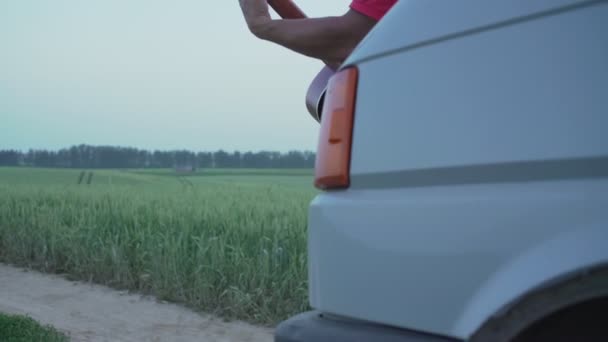 Man in rode trui met baard in tarweveld speelt akoestische gitaar op kap van auto — Stockvideo