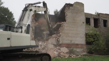 Büyük demir kovalı kazıcı, eski beton bina duvarını yıkıyor.