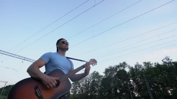 Músico con guitarra usando gafas de sol y gorra va a lo largo del ferrocarril — Vídeo de stock