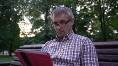 Adam şehir parkında otururken tablet kullanıyor.