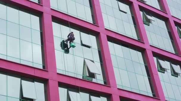 Рабочий в шлеме висит на веревке на высоте и моет окна стекла — стоковое видео