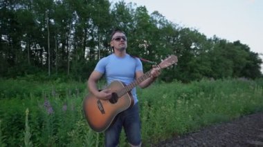 Demiryolunda güneş gözlüğü ve şapka takan erkek müzisyen gitar çalar ve şarkı söyler.