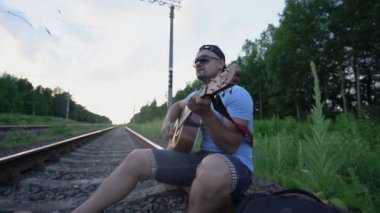 Güneş gözlüklü ciddi bir adam demiryolunda oturur gitar çalar, şarkı söyler ve başka yere bakar.