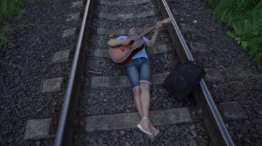 Güneş gözlüklü ve şapkalı bir adam demiryollarında yatıyor gitar çalıyor ve şarkı söylüyor.